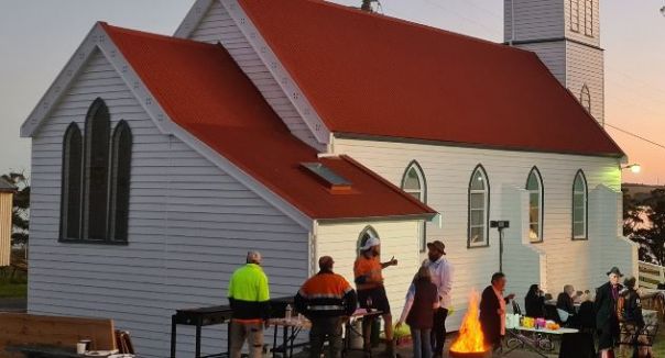 The Church can been seen in this image after it has had renovations. The community can be seen celebrating outside of the church, drinking and eating. The sun shines on the side of the Church. 