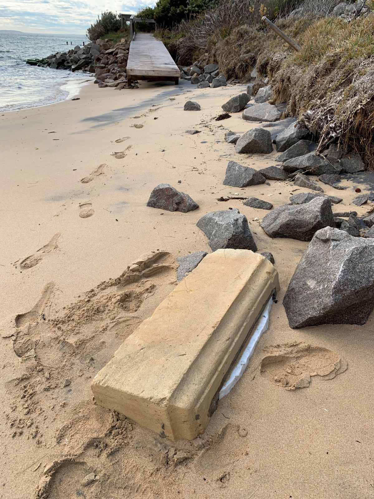 A broken tombstone can be seen nested in the sand by rocks. The ocean can be seen in the foreground.